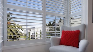 A room with a grey armchair and a red pillow, a white ottoman, and a window with white blinds.