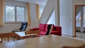 A room with a dining table, two chairs, a red sofa, and a black chair. There is a window with curtains, a door leading to another room, and a white wall with a triangular architectural feature. - 2-room vacation apartment for 4 people on the 1st floor - with WLAN - NH (Ribnitz-Damgarten)