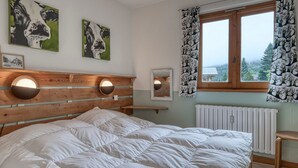 A bedroom with a wooden headboard, two wall-mounted lights, a bed with white bedding, a window with patterned curtains, and two paintings on the wall.