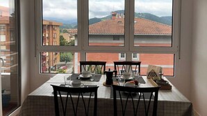 A dining area with a table set for four, large windows with a view of buildings and hills, and a ceiling light fixture. - Piso Céntrico y Tranquilo en Gernika (Gernika-Lumo)