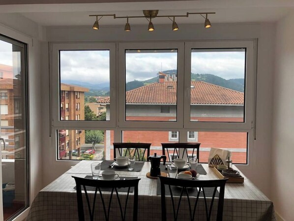 Comedor con una mesa dispuesta para cuatro personas, ventanas amplias con vista a edificios y colinas, y una lámpara en el techo.