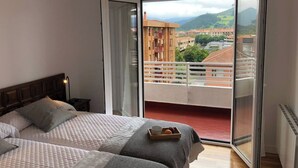 A bedroom with two beds, a balcony with a view of buildings and mountains, and a cityscape visible through the window. - Piso Céntrico y Tranquilo en Gernika (Gernika-Lumo)