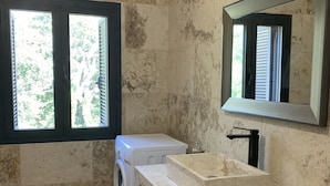 Une salle de bain avec un sol en marbre, un lave-linge, un lavabo avec un miroir et une fenêtre avec des stores.