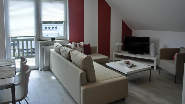 A modern living room with a sofa, coffee table, and TV. There is a window with blinds and a view of the outside.