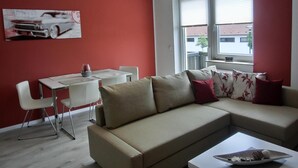 A modern living room with a beige sofa, a dining table with chairs, and a large window with blinds.