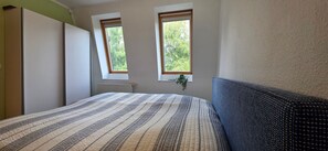 Ein Schlafzimmer mit einem Bett, zwei Fenstern, einem Heizkörper und einer Pflanze.