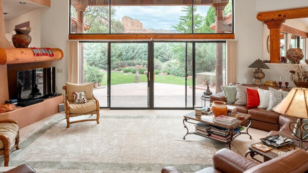 A spacious living room with a fireplace, comfortable seating, and large windows offering a view of a garden and mountains. - Gated Historic Estate • Pool, Spa & Artifacts (Sedona)