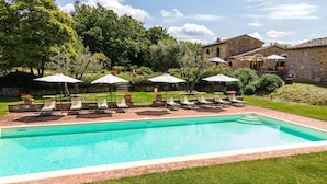 Une piscine entourée d’un bâtiment en pierre et d’une végétation luxuriante, avec des chaises longues et des parasols à proximité.
