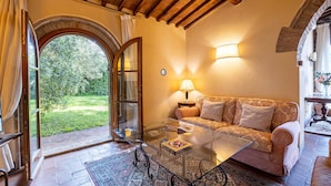 Un salon chaleureux avec une table basse en verre, un canapé agrémenté de coussins à motifs, une lampe et une vue sur un jardin par une porte ouverte.