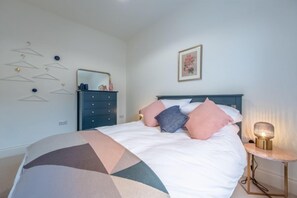 Cama bem arrumada com manta de padrão geométrico, mesa de cabeceira com abajur e cômoda com espelho.