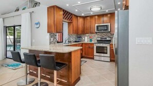 Une cuisine moderne avec des meubles en bois, un réfrigérateur en acier inoxydable, un four à micro-ondes et un four traditionnel. On y trouve également un coin bar avec deux chaises et un petit îlot central doté d’un évier.
