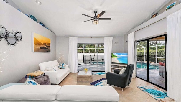 Un salon moderne avec un canapé blanc, un fauteuil noir et une table basse. On y trouve un ventilateur de plafond, un tableau au mur et, à travers de grandes portes vitrées, une vue sur une piscine.