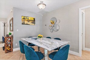Een eetgedeelte met een marmeren tafel, blauwe stoelen, een vaas met gele bloemen en een schilderij aan de muur.
