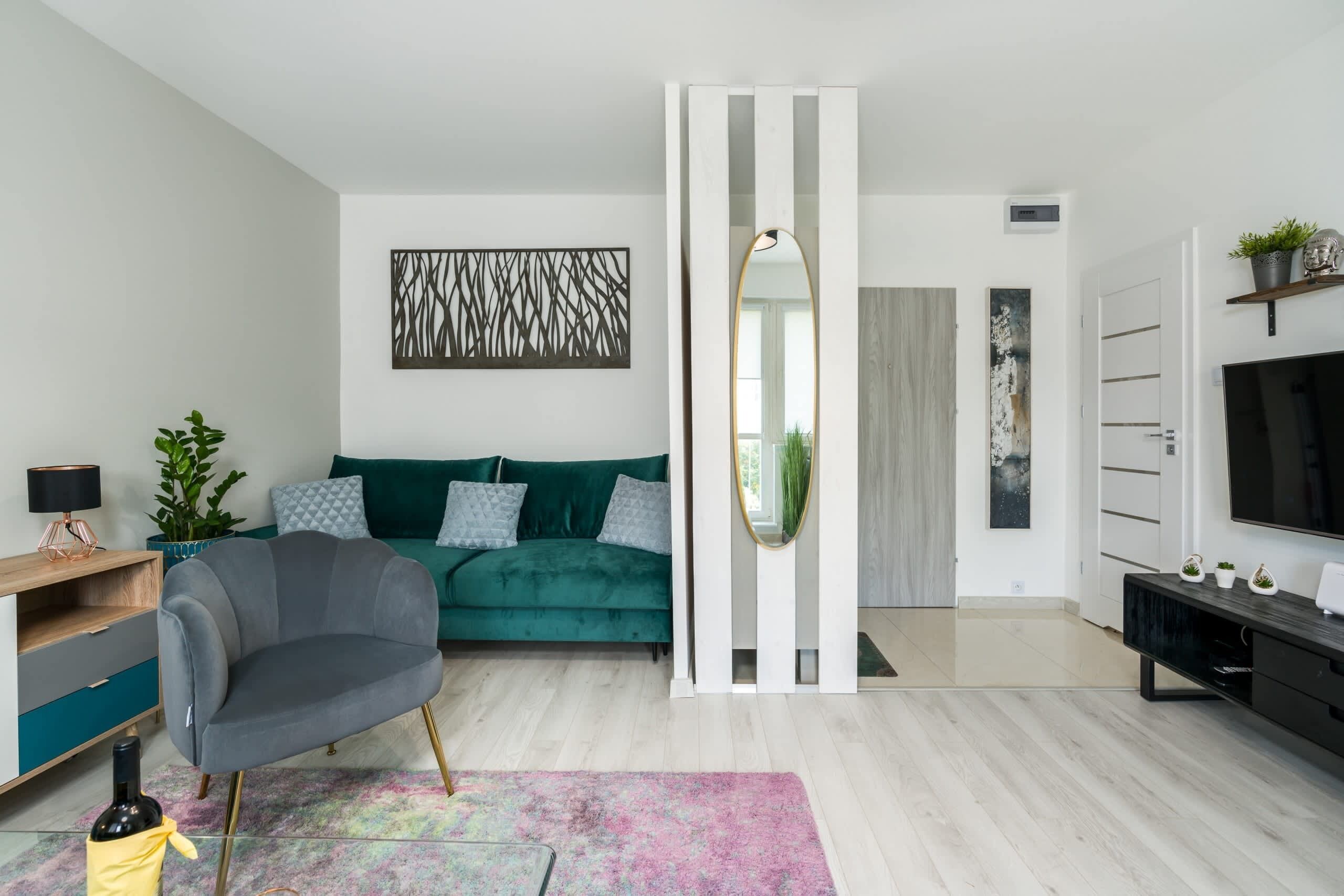 Una sala de estar moderna con un sofá azul verdoso, un sillón gris, un espejo grande y un televisor sobre un soporte negro.