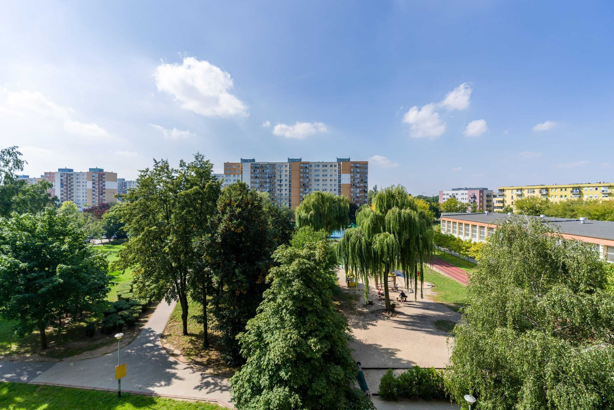 Ein Wohngebiet mit mehrstöckigen Wohngebäuden, einem Park mit Bäumen und einer Laufbahn, und einem strahlend blauen Himmel.