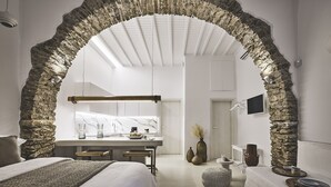 A modern bedroom with a stone archway, a large bed, a bedside table, a desk with a chair, a small round ottoman, and a white sofa. - Crystal Green Signature Suites Mykonos (Mykonos)