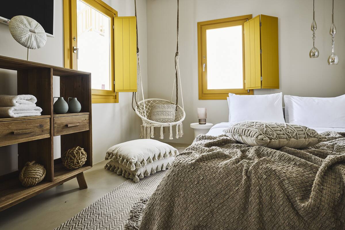 A bedroom with a bed, a hanging chair, a wooden shelf with towels and vases, and a window with yellow shutters.