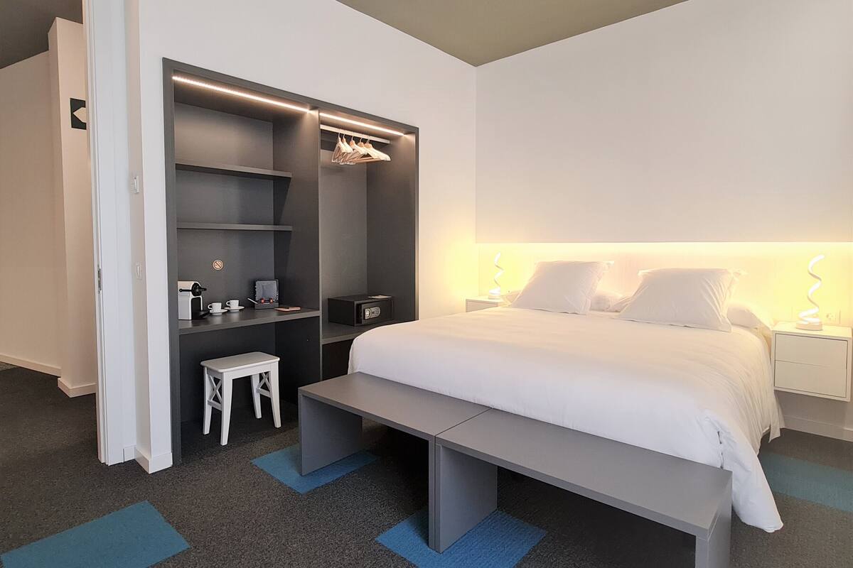 Una habitación de hotel moderna con una cama, un escritorio, una silla, un taburete pequeño y un armario.