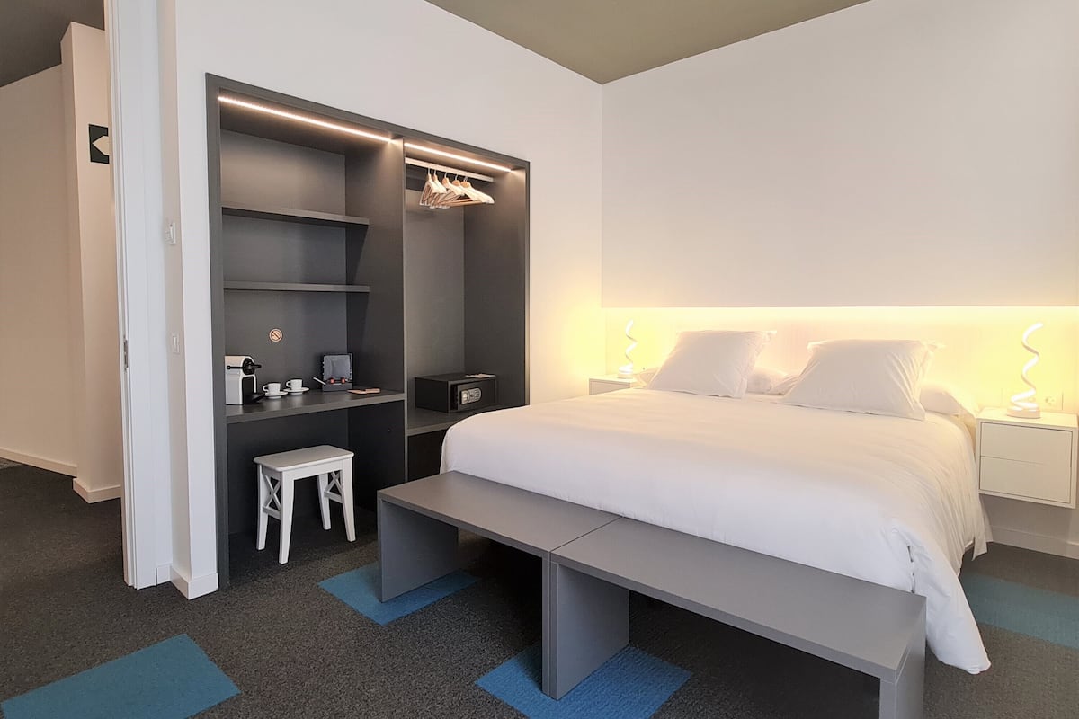 Una habitación de hotel moderna con una cama, mesita de noche, un escritorio con silla, un armario y un taburete pequeño.