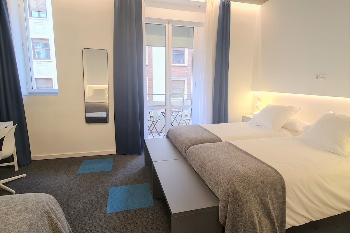Habitación de hotel con una cama grande, una mesita de noche, una silla, un escritorio pequeño, un espejo y una ventana con cortinas.