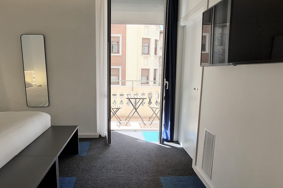 Habitación de hotel con cama, mesita de noche, espejo, televisor y balcón con vistas a edificios.