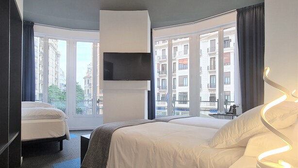 Quarto de hotel moderno com uma cama grande, uma TV fixada na parede e vista para a cidade através de amplas janelas.