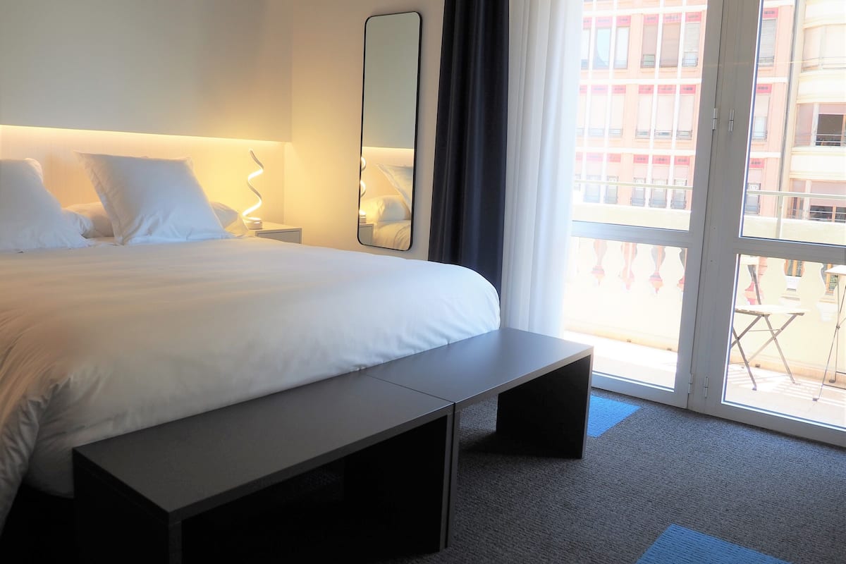 Habitación de hotel con una cama grande, una mesita de noche, un espejo y vistas al balcón exterior.