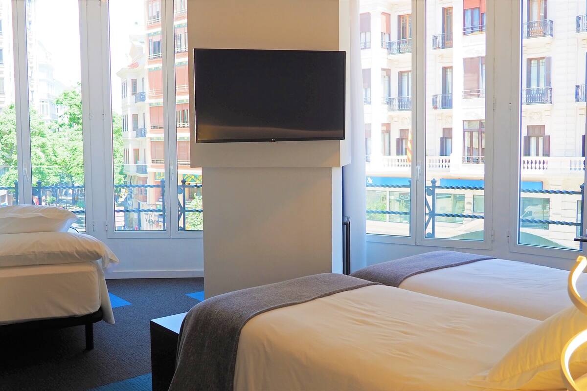 Habitación de hotel con dos camas, un televisor en la pared y vistas a un paisaje urbano a través de amplias ventanas.