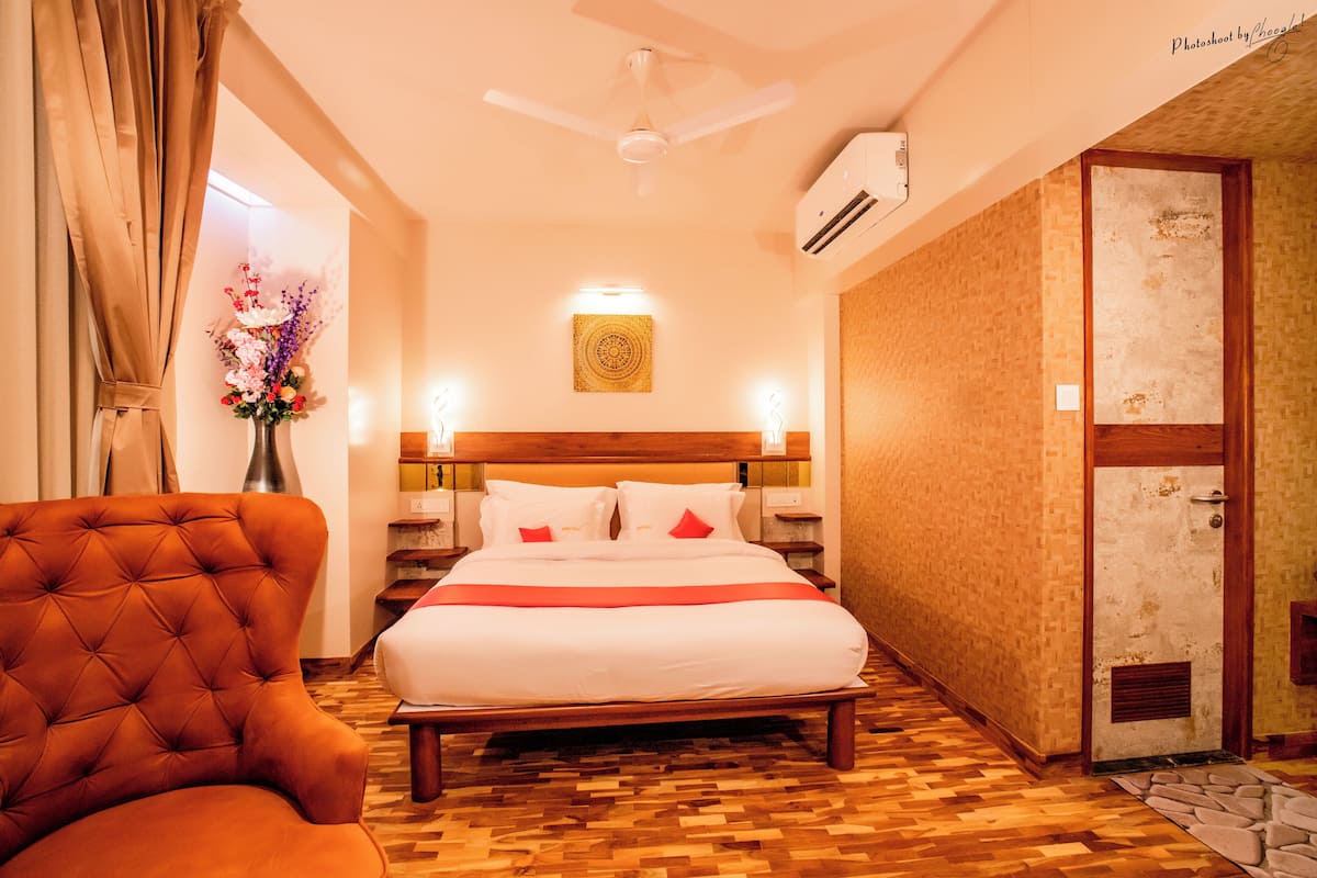 Quarto de hotel com uma cama grande, uma poltrona estofada, um ventilador de teto e um ar condicionado fixado na parede.