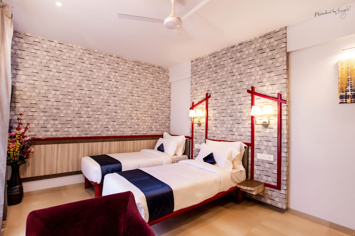 Quarto de hotel com duas camas, mesa de cabeceira com moldura vermelha, um vaso com flores e uma luminária fixada na parede.