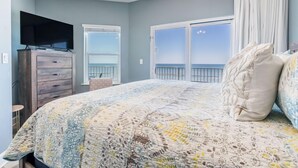 Ein Schlafzimmer mit einem großen Bett, einem Schminktisch, einem Fernseher darauf und Blick auf das Meer durch die Fenster.