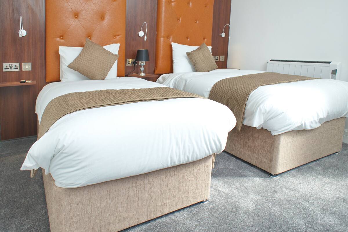 Two beds with headboards, a bedside table with a lamp, and a wall-mounted thermostat.