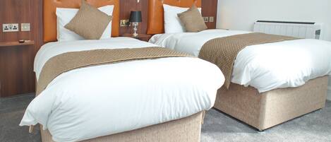 Two beds with headboards, a bedside table with a lamp, and a wall-mounted thermostat.