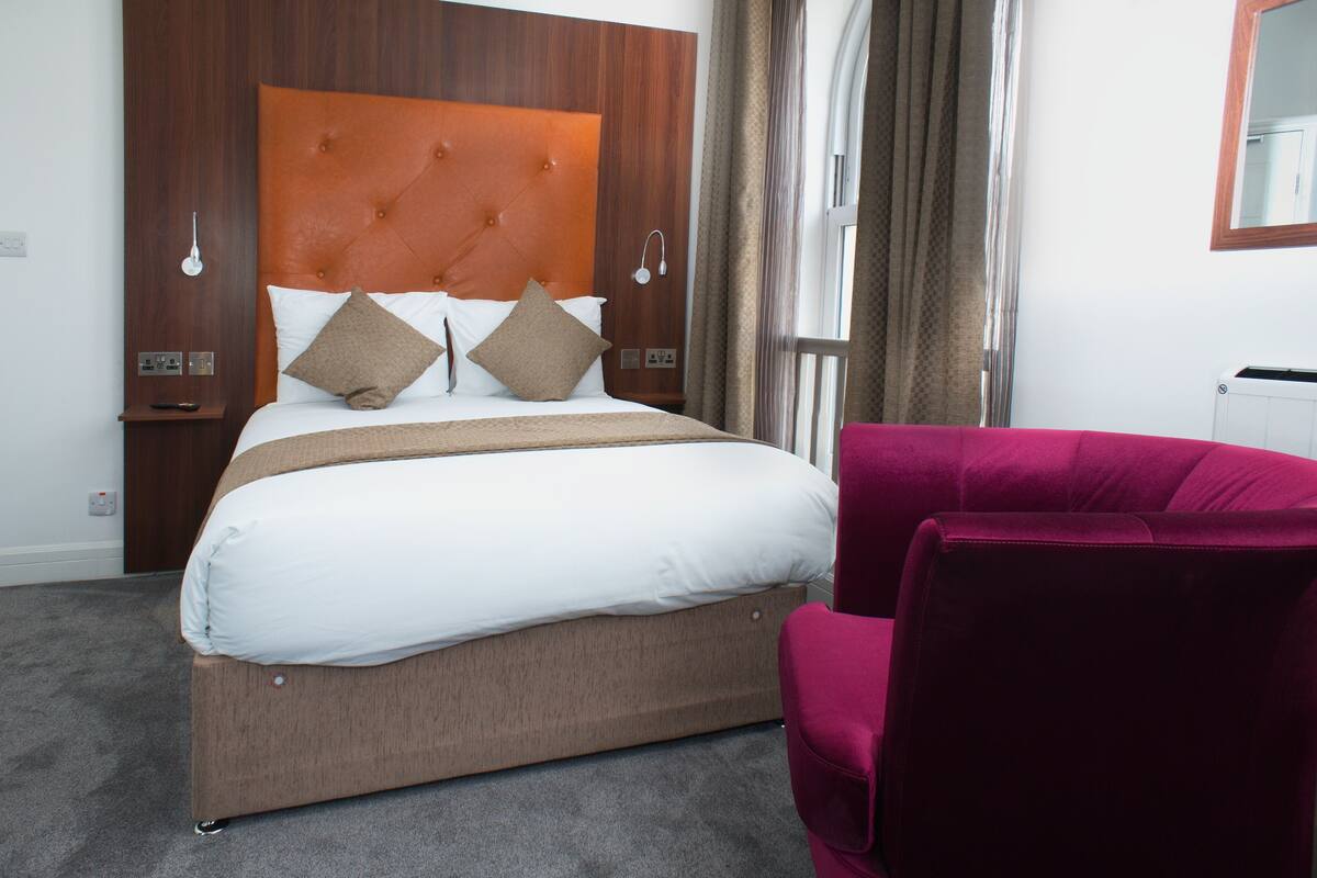 A hotel room with a large bed, a wooden headboard, a red armchair, and a window with curtains.
