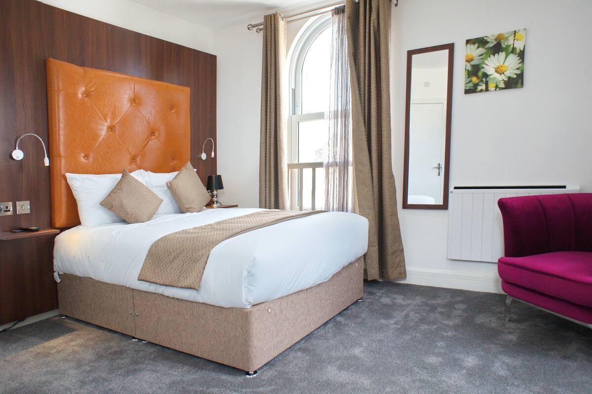 A hotel room with a large bed, a wooden headboard, a nightstand, a lamp, a window with curtains, a radiator, a mirror, and a pink armchair.