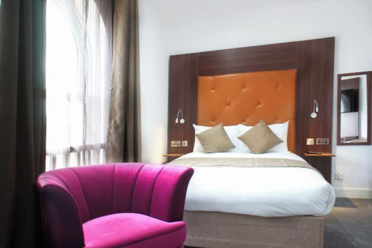 A hotel room with a large bed, a wooden headboard, two bedside tables with lamps, and a pink armchair.