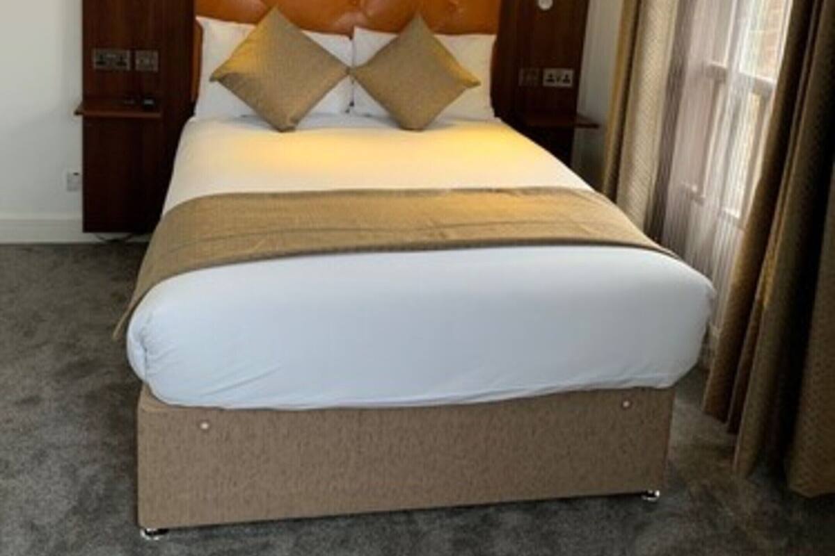 A hotel room with a large bed, wooden headboard, two bedside tables with lamps, and a view of the outdoors through a window with curtains.
