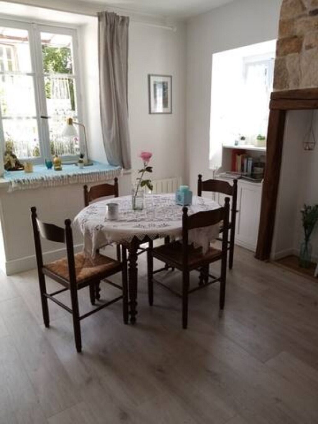A dining area with a round table, four chairs, a vase with a single flower, and a window with curtains.