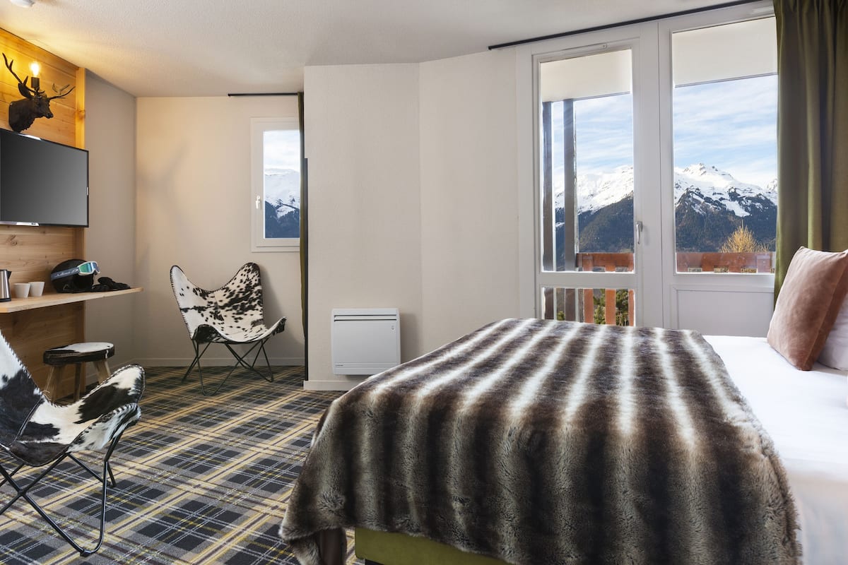 Habitación de hotel con cama, una silla, un escritorio, televisor y ventana con vista a la montaña.