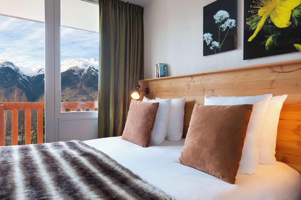 A bed with a view of mountains through a window, a wooden headboard, and a painting of flowers.