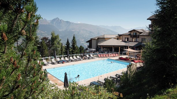 Una piscina con sillones de descanso, un edificio con balcón y montañas al fondo.