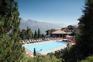 Une piscine avec des chaises longues, un bâtiment doté d’un balcon et des montagnes en arrière-plan.