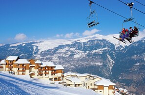 A ski resort with multiple buildings, a ski lift, and snow-covered mountains in the background.