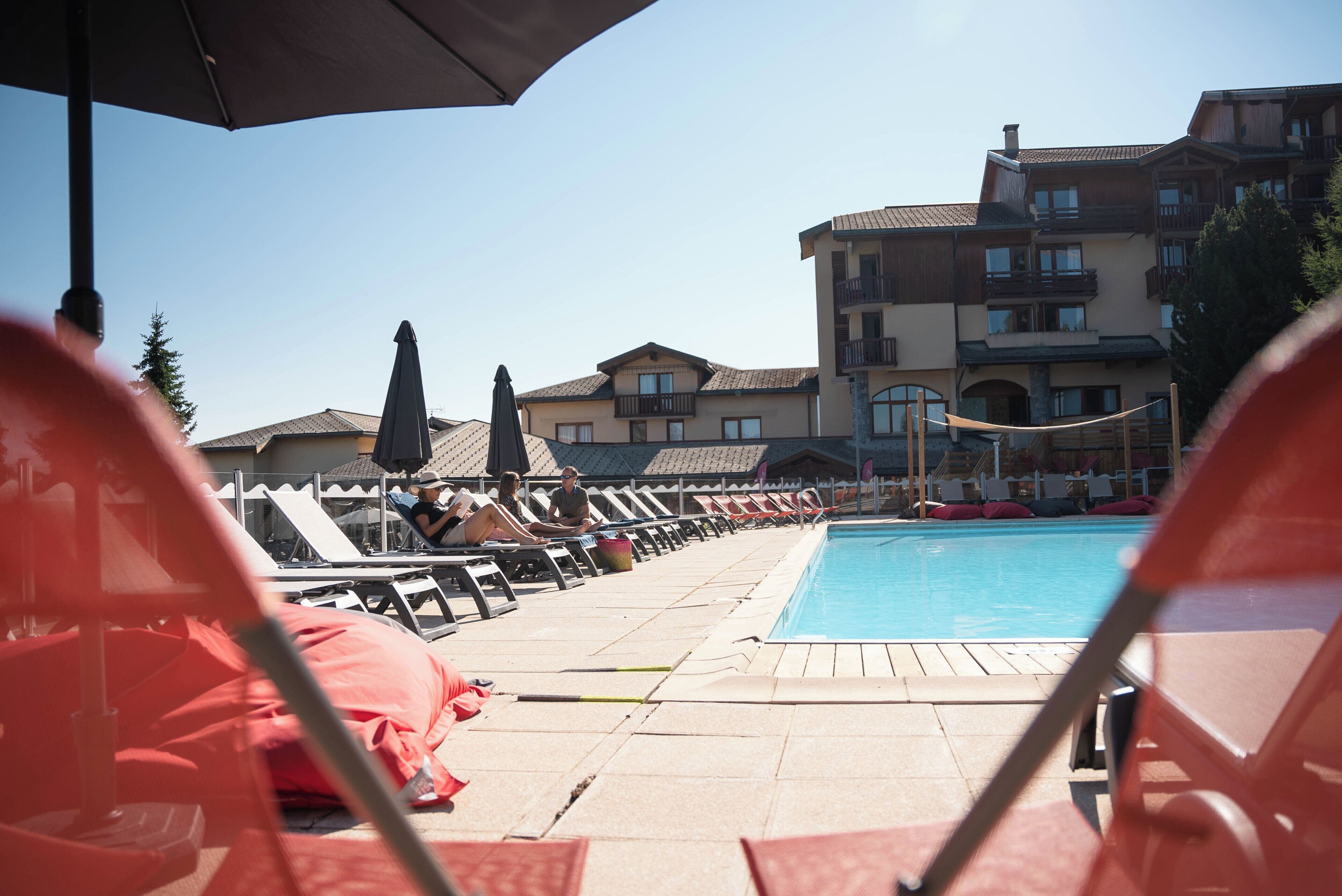 A poolside area with lounge chairs and umbrellas, a swimming pool, and a building with balconies in the background.