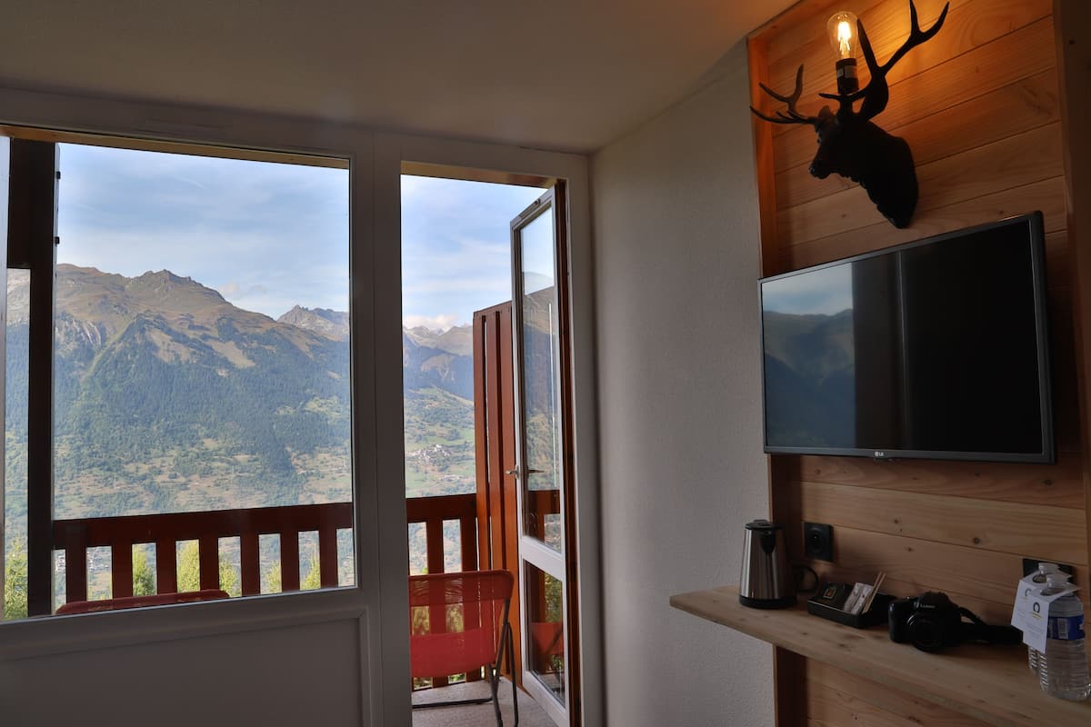 Una habitación con una pared de madera y un televisor fijado, una cabeza de ciervo montada en la pared, una silla roja y vistas a montañas a través de un ventanal.