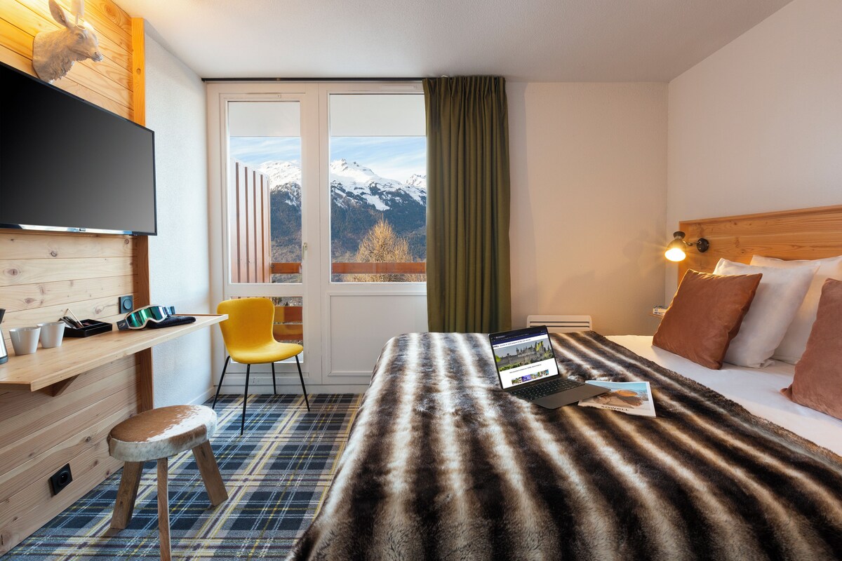 Habitación de hotel con cama, escritorio, silla, televisión y ventana con vista a la montaña.