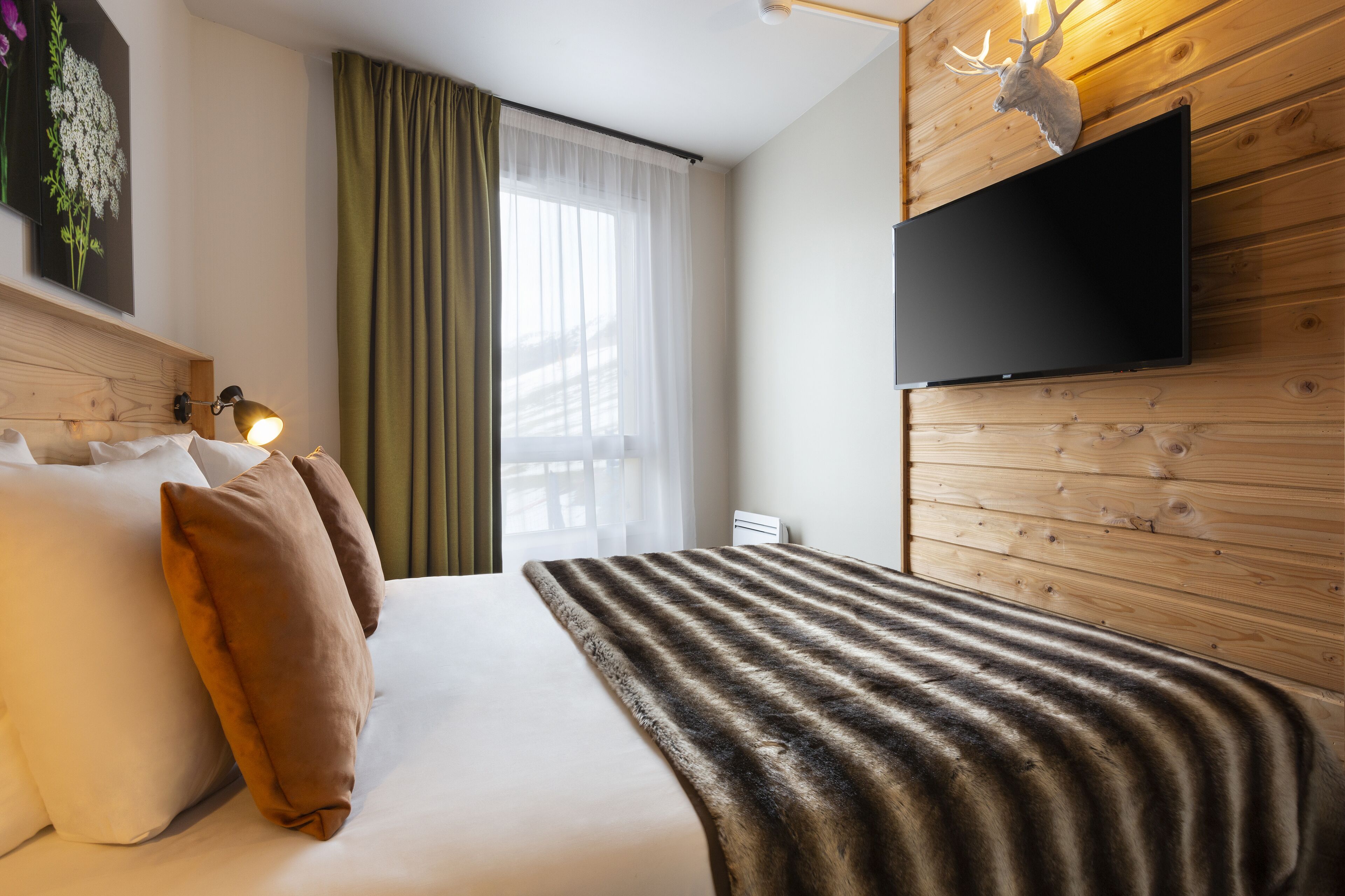 A hotel room with a bed, pillows, a blanket, a television mounted on a wooden wall, and a window with curtains.