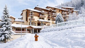 Un hotel de varios pisos con balcones, una entrada cubierta de nieve y una pista de esquí al fondo.