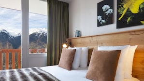 Una cama con vistas a montañas a través de una ventana, cabecera de madera y un cuadro de flores.