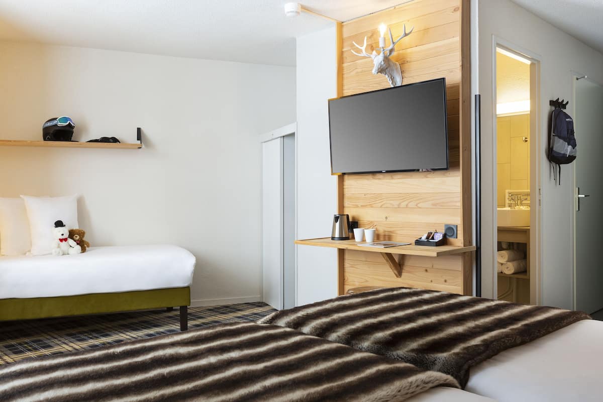 Habitación de hotel con cama, televisor montado en la pared, estante de madera con un casco y un oso de peluche, y una puerta que da a un baño.